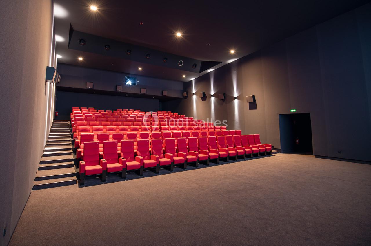 Salle de cinéma vide avec rangées de fauteuils rouges, éclairage tamisé et écran éteint en arrière-plan.