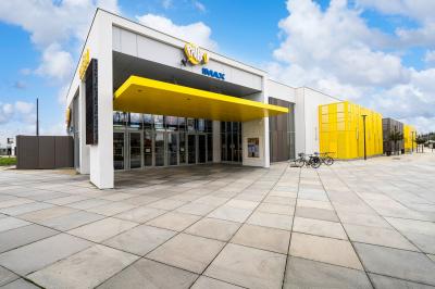 Façade d'un cinéma moderne avec enseigne IMAX, éclairée en soirée, entourée de végétation et d'un parking.