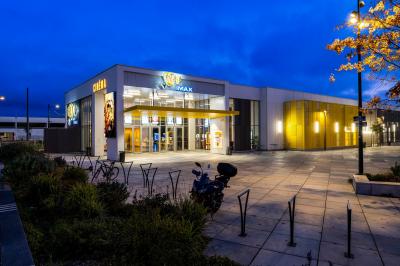 Façade d'un cinéma moderne avec enseigne IMAX, éclairée en soirée, entourée de végétation et d'un parking.