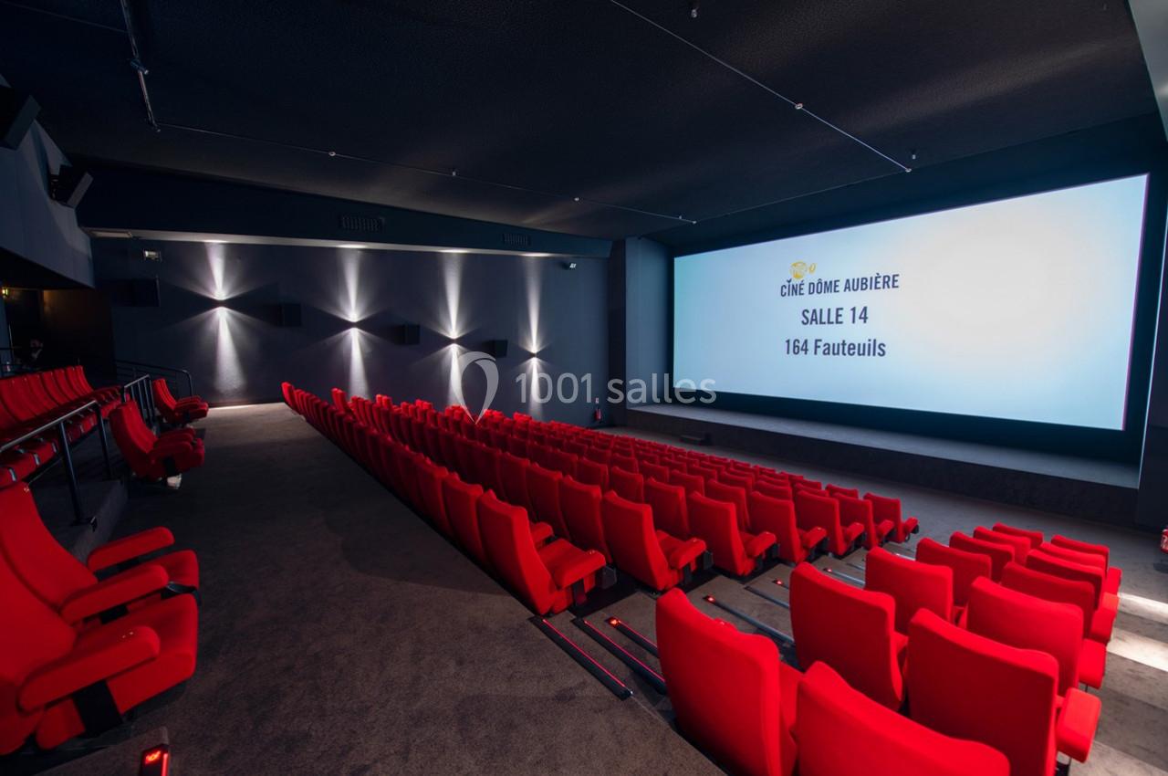 Salle de cinéma avec écran allumé, rangées de fauteuils rouges et éclairage tamisé.