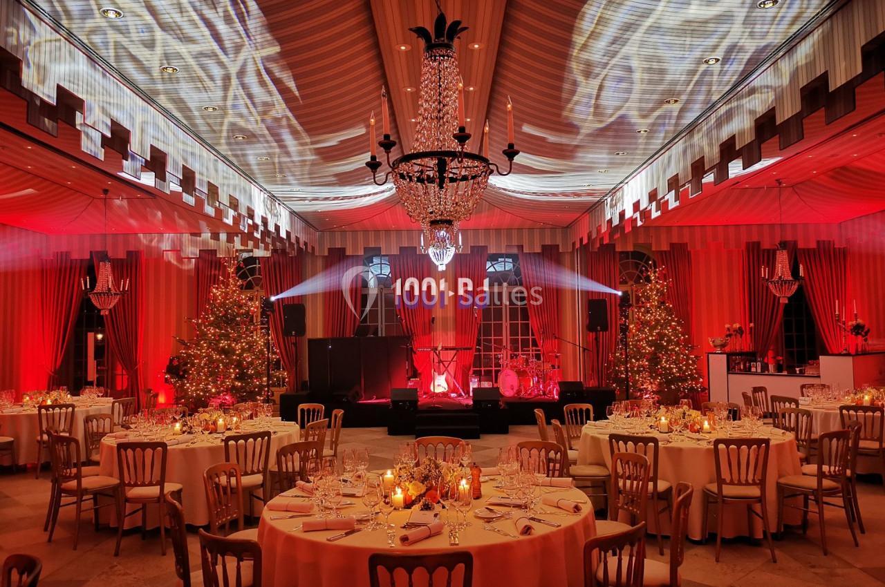 Salle de réception décorée avec des tables dressées, éclairage rouge, sapins de Noël et un lustre central.