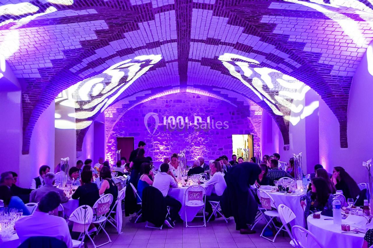 Salle voûtée en briques éclairée par des lumières violettes, avec des invités assis à des tables lors d'un événement.