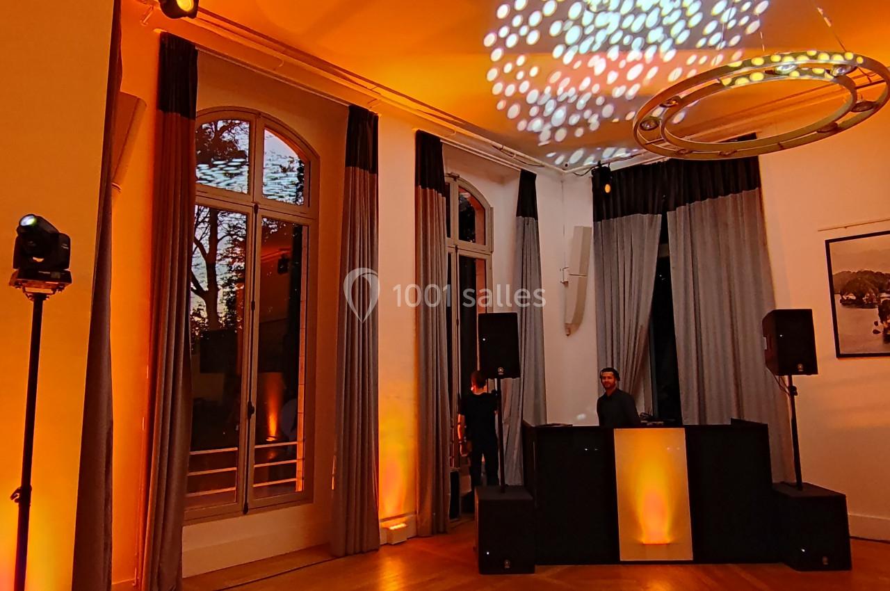 Salle éclairée en orange avec un DJ derrière une table, des enceintes et des projections lumineuses au plafond.