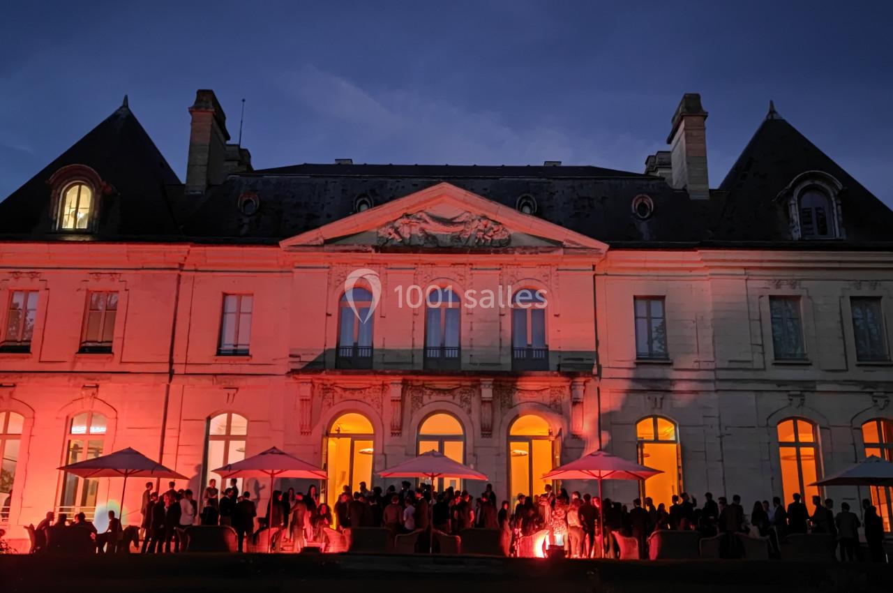 Façade d'un château éclairée en rouge à la tombée de la nuit, avec des invités rassemblés sous des parasols.