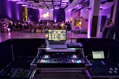 Vue d'une régie DJ avec platines et ordinateur, face à une salle éclairée en violet lors d'un événement festif.