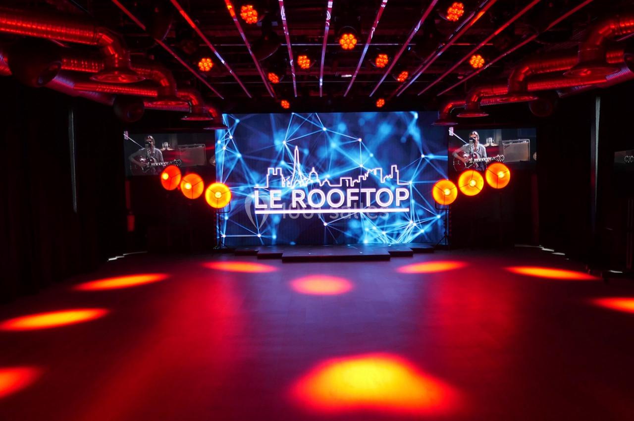 Salle avec un écran géant affichant ’Le Rooftop’, éclairée par des projecteurs rouges et un plafond avec des structures…