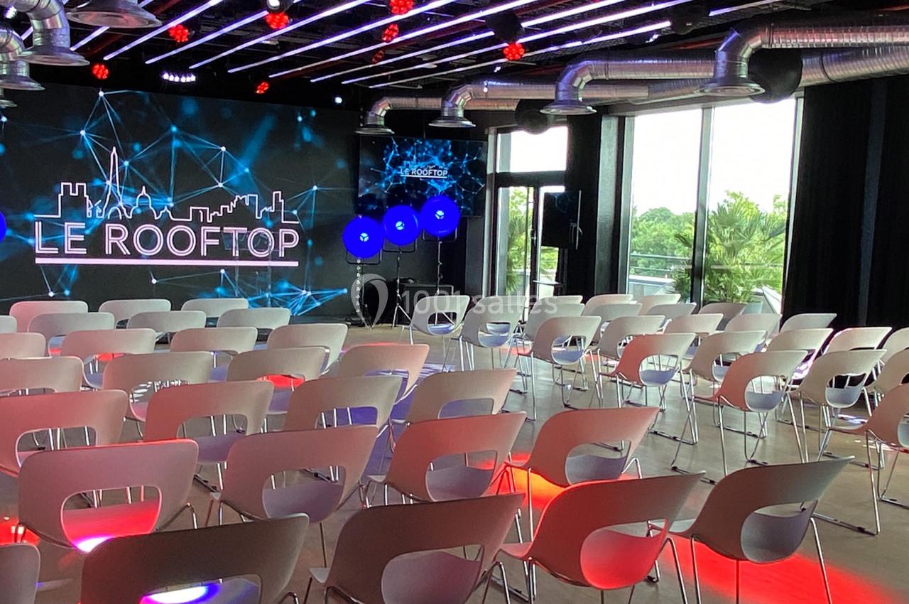 Salle de conférence moderne avec chaises blanches alignées, écran géant et éclairage coloré, vue sur l'extérieur.