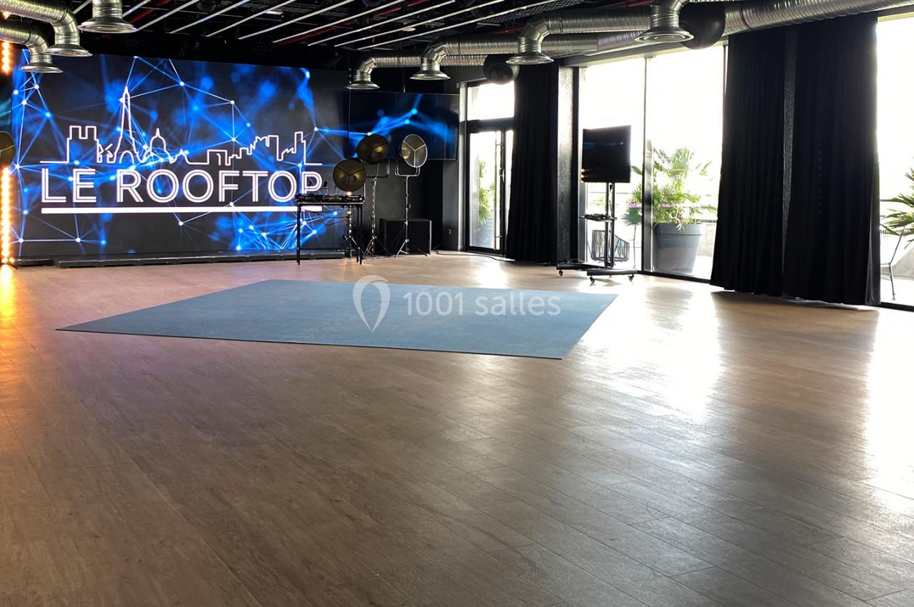 Salle lumineuse avec parquet, grande baie vitrée, écran LED affichant ’Le Rooftop’ et projecteurs en arrière-plan.