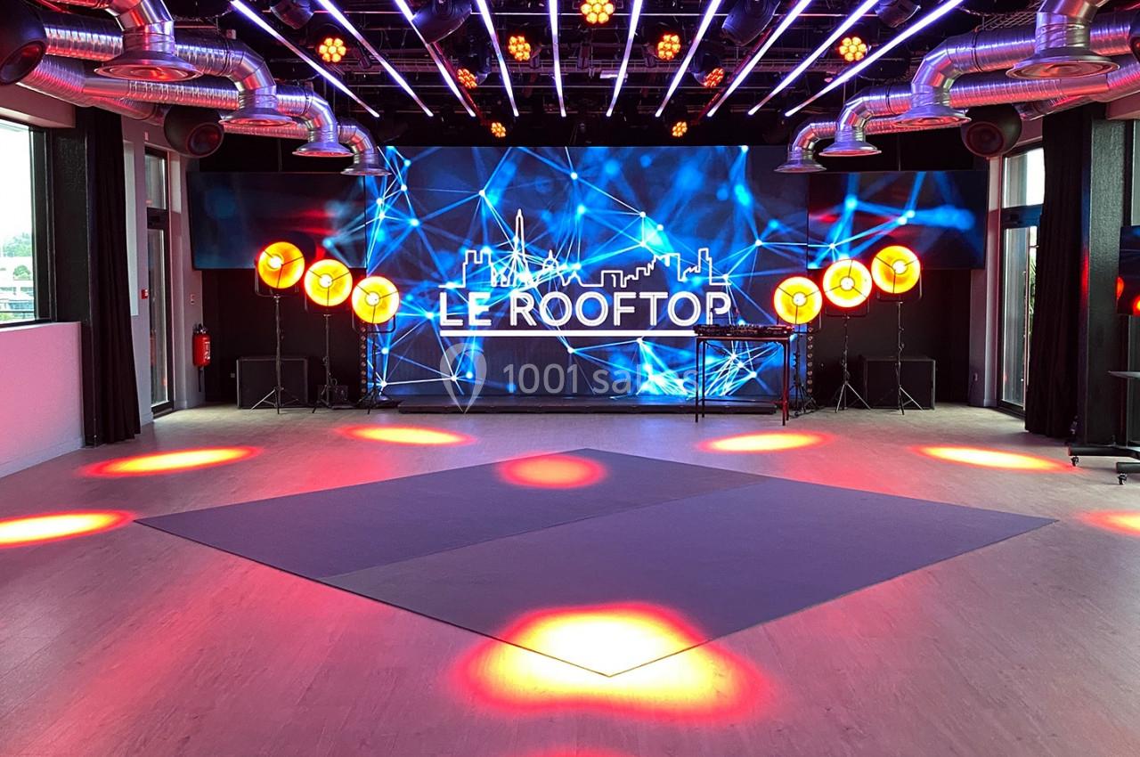 Salle lumineuse avec écran géant affichant ’Le Rooftop’, éclairages colorés et grandes baies vitrées.
