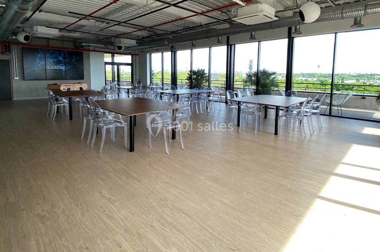 Salle lumineuse avec de grandes baies vitrées, tables en bois, chaises transparentes et vue sur un espace extérieur…