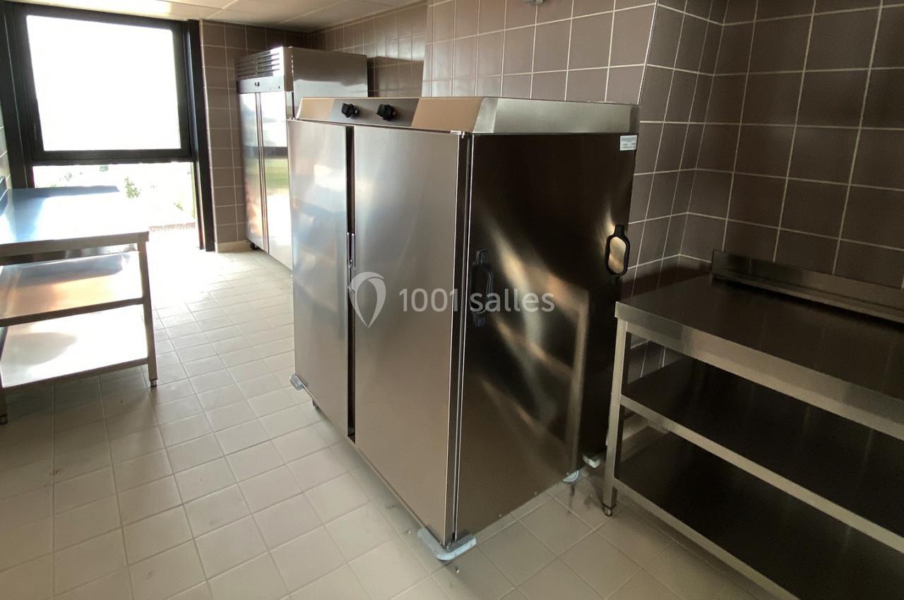 Cuisine professionnelle avec réfrigérateur en inox, étagères métalliques et plan de travail, éclairée par une fenêtre.