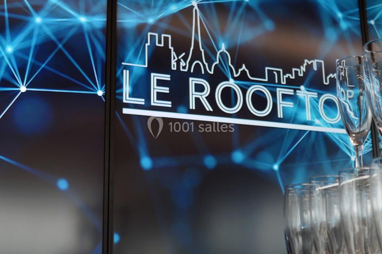 Des verres à champagne empilés devant un écran affichant un logo ’Le Rooftop’ avec un fond de motifs lumineux bleus.