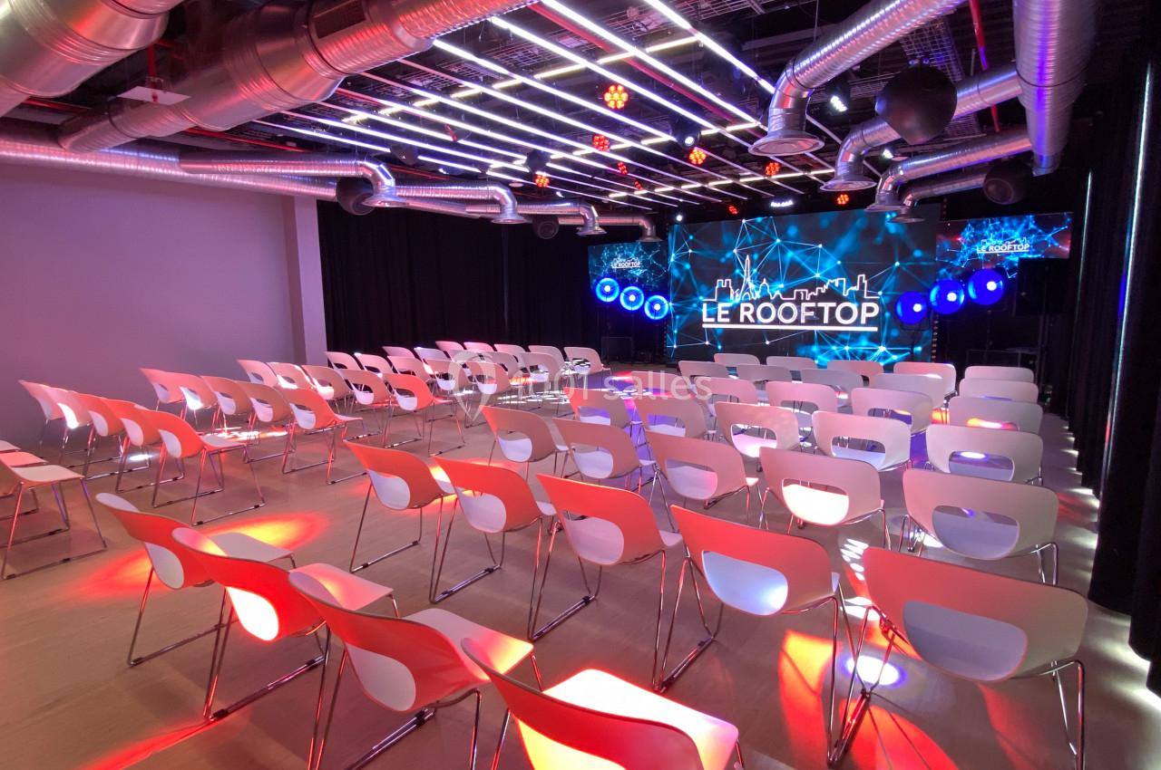 Salle moderne avec rangées de chaises blanches, éclairage coloré et écran LED affichant ’Le Rooftop’.