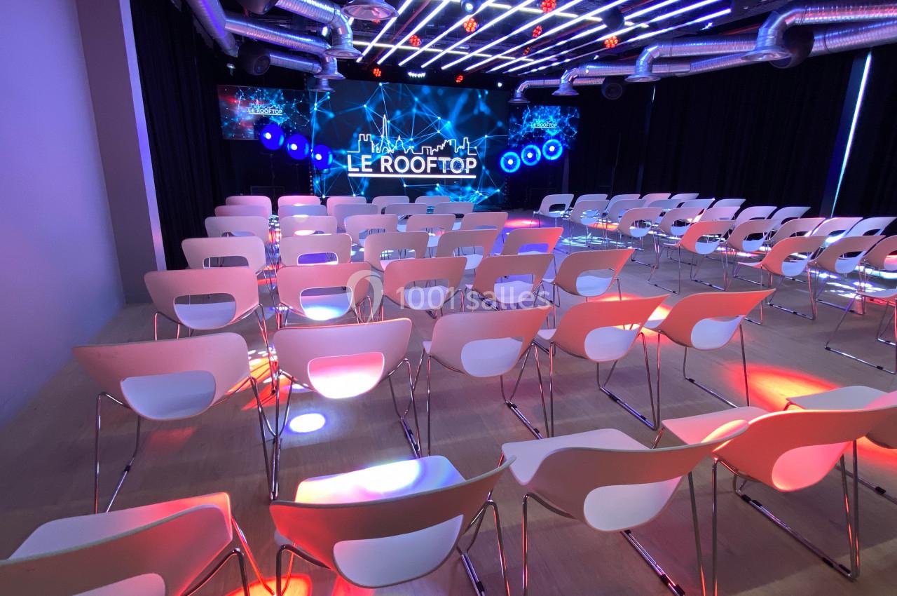 Salle de conférence moderne avec rangées de chaises blanches, éclairage coloré et écran affichant ’Le Rooftop’.