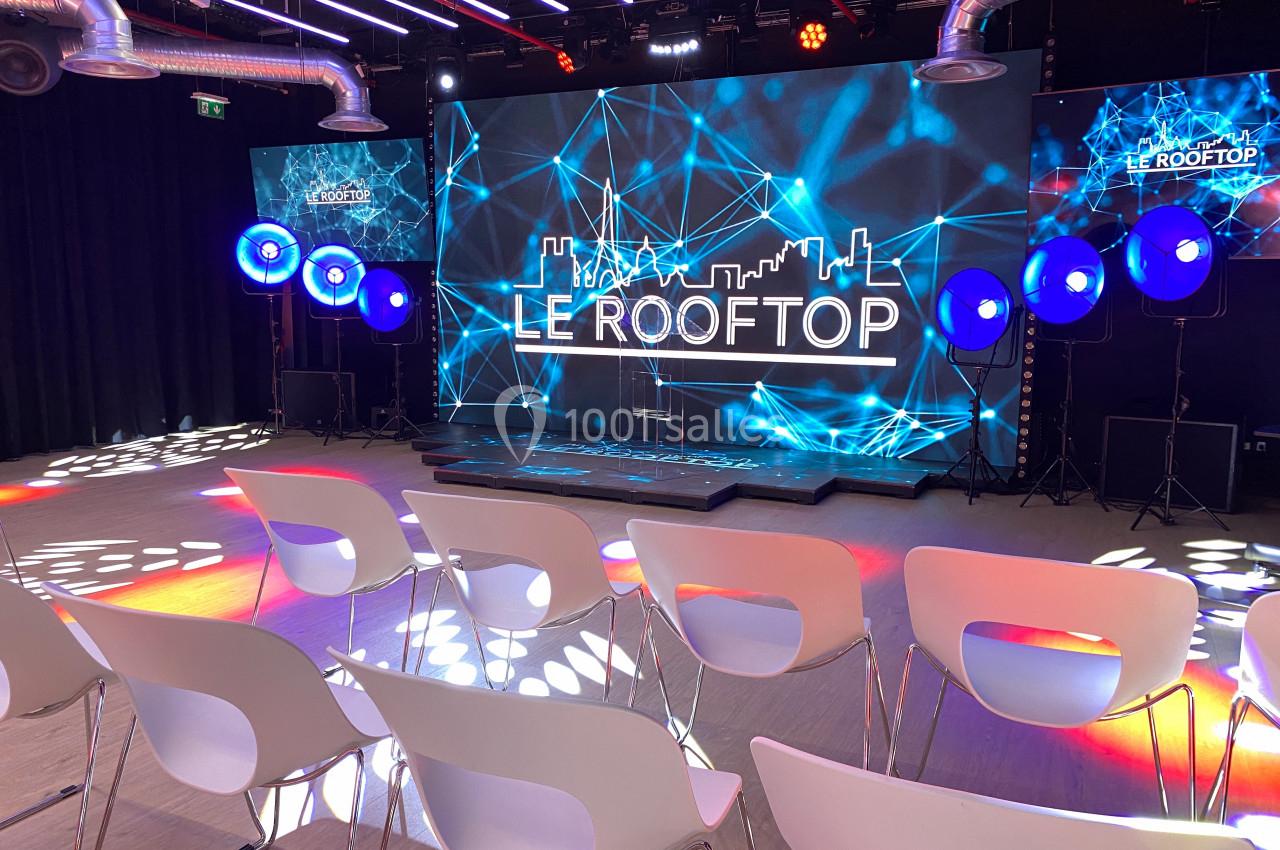 Salle de conférence moderne avec scène éclairée, écrans LED affichant ’Le Rooftop’ et rangées de chaises blanches.