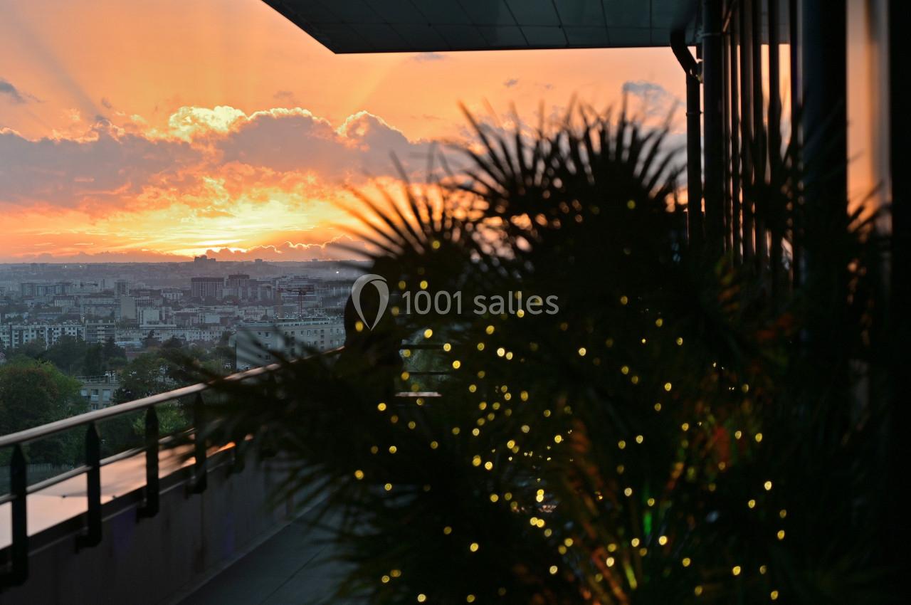 Coucher de soleil sur une ville, vu depuis un balcon avec des plantes illuminées au premier plan.