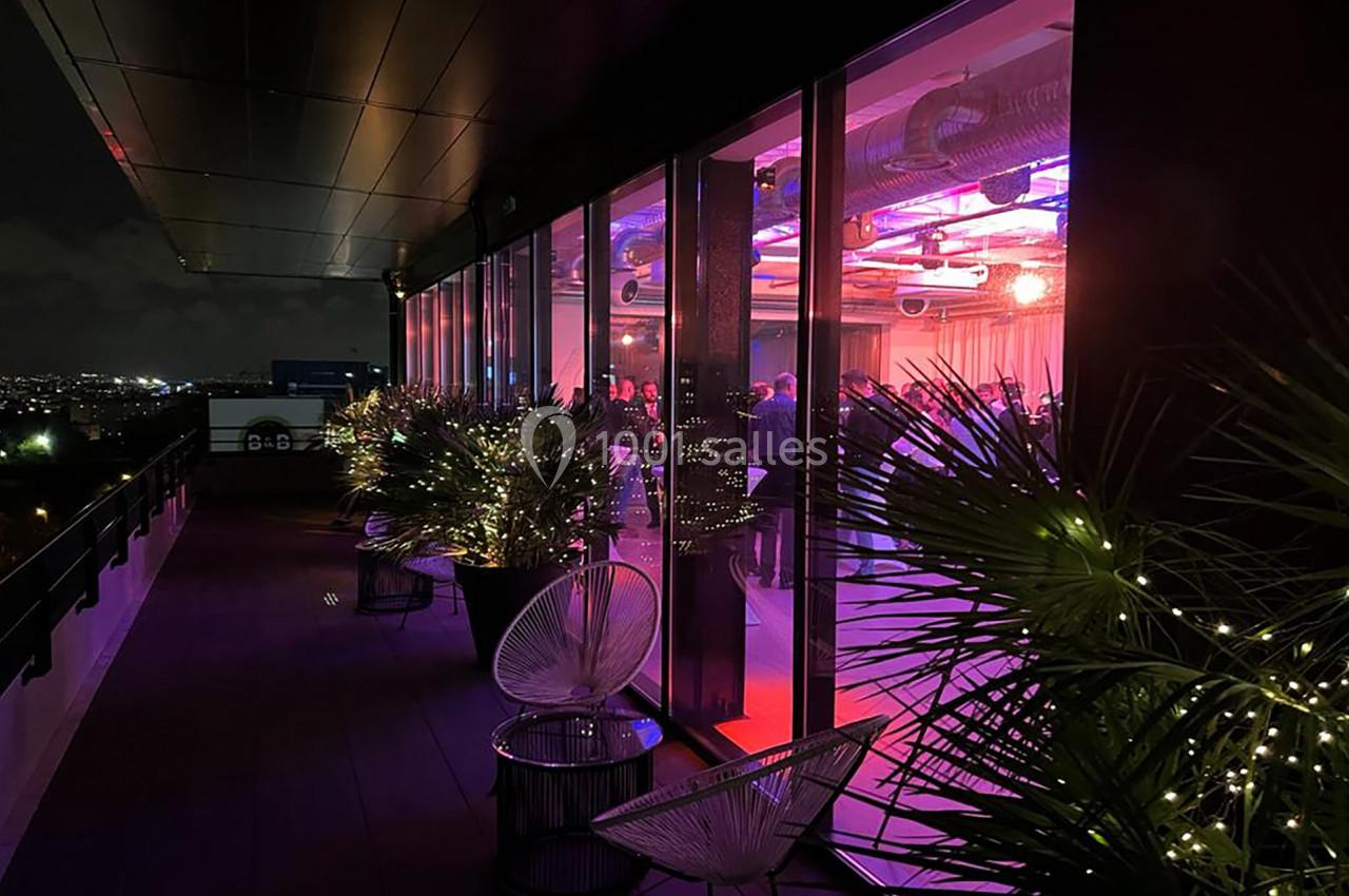 Vue nocturne d'une terrasse illuminée avec des plantes et des fauteuils, donnant sur une salle éclairée aux tons colorés.
