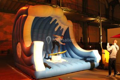 Miniature ArScénique #13 Homme sur une planche de surf simulant une vague géante gonflable dans un espace intérieur éclairé.