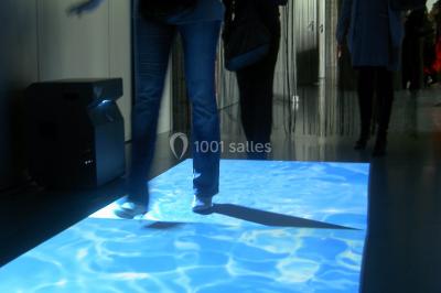 Miniature ArScénique #3 Homme sur une planche de surf simulant une vague géante gonflable dans un espace intérieur éclairé.