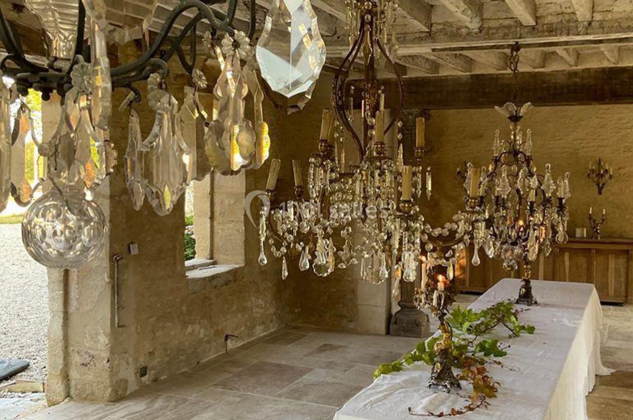 Salle rustique avec des lustres en cristal suspendus, tables recouvertes de nappes blanches et décoration végétale.