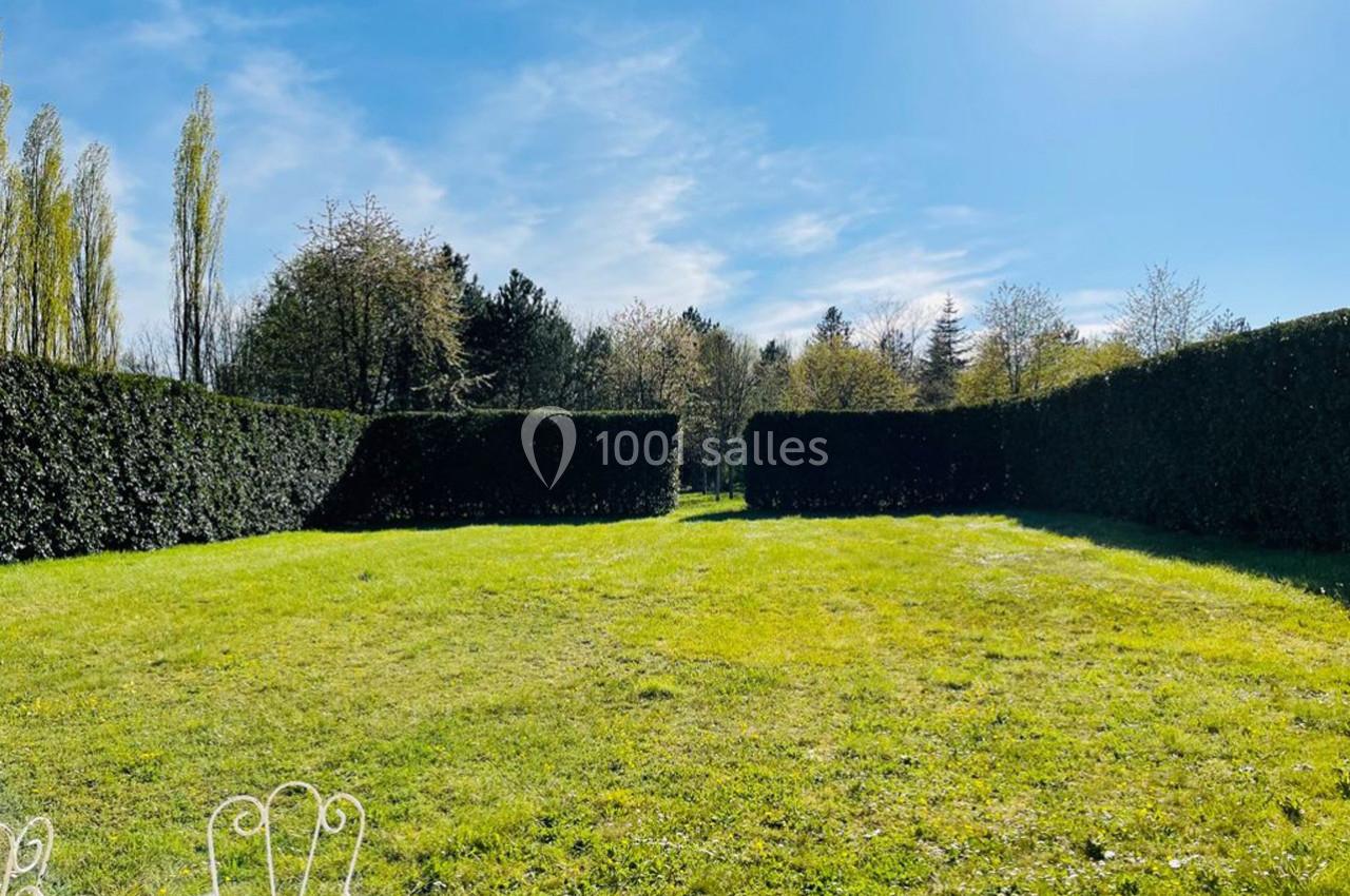 Pelouse entourée de haies taillées, avec des arbres en arrière-plan sous un ciel bleu ensoleillé.