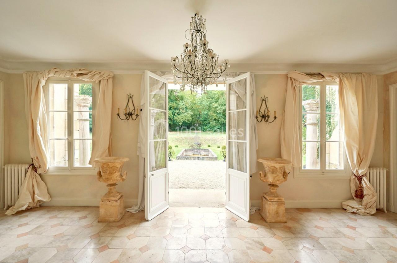 Salle lumineuse avec portes-fenêtres ouvertes sur un jardin, lustre central et décorations en pierre.