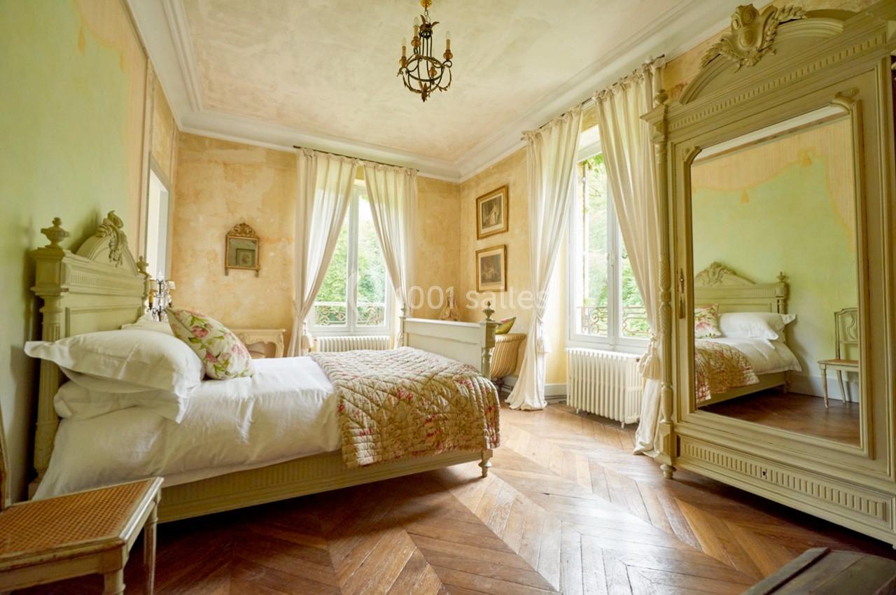 Chambre lumineuse avec lit ancien, parquet en bois, miroir orné et grandes fenêtres donnant sur un jardin.