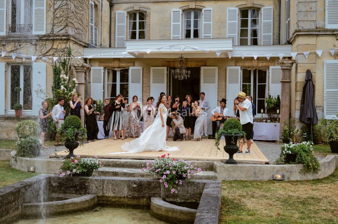 Un couple danse sur une estrade en bois devant un manoir, entouré d'invités applaudissant.