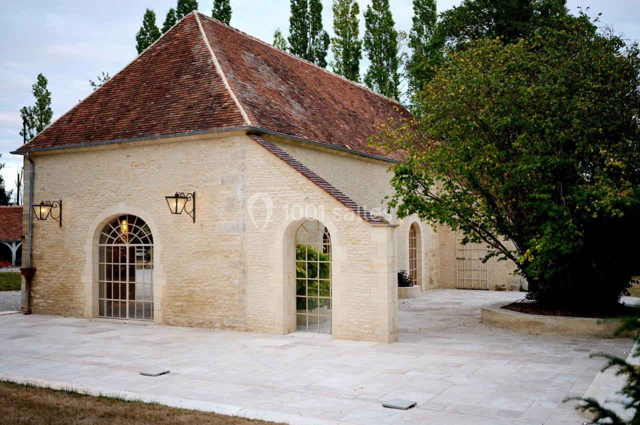 Bâtiment en pierre avec toit en tuiles rouges, grandes fenêtres en arc et terrasse pavée entourée de verdure.