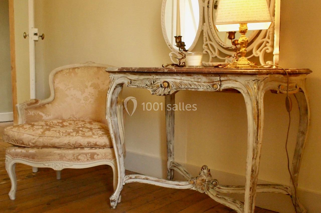 Fauteuil et console en bois sculpté de style ancien, avec une lampe allumée et un miroir, dans une pièce au sol en bois.