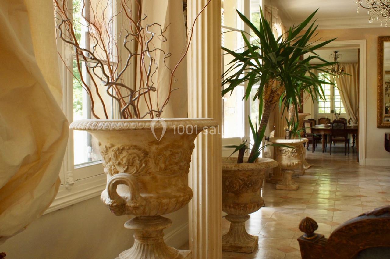 Grande pièce lumineuse avec des colonnes, des plantes en pot et des urnes décoratives en pierre près des fenêtres.