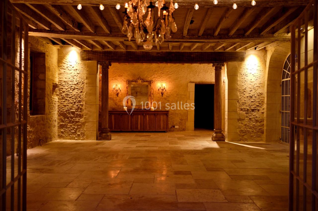 Salle en pierre avec un sol en dalles, un lustre suspendu, des murs éclairés et un meuble en bois au fond.