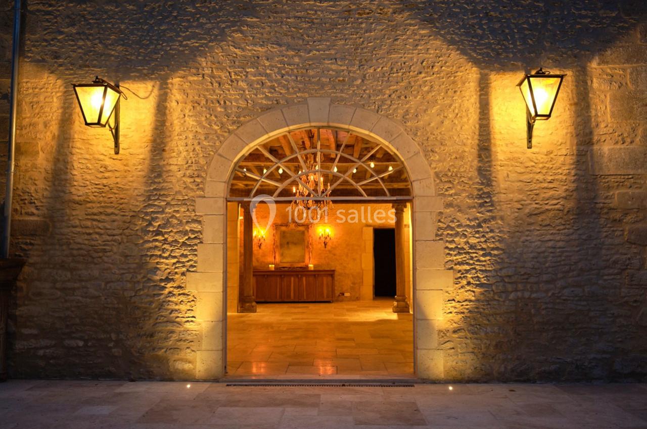 Entrée voûtée en pierre éclairée par des lanternes, donnant sur une salle avec un lustre et des murs en pierre.