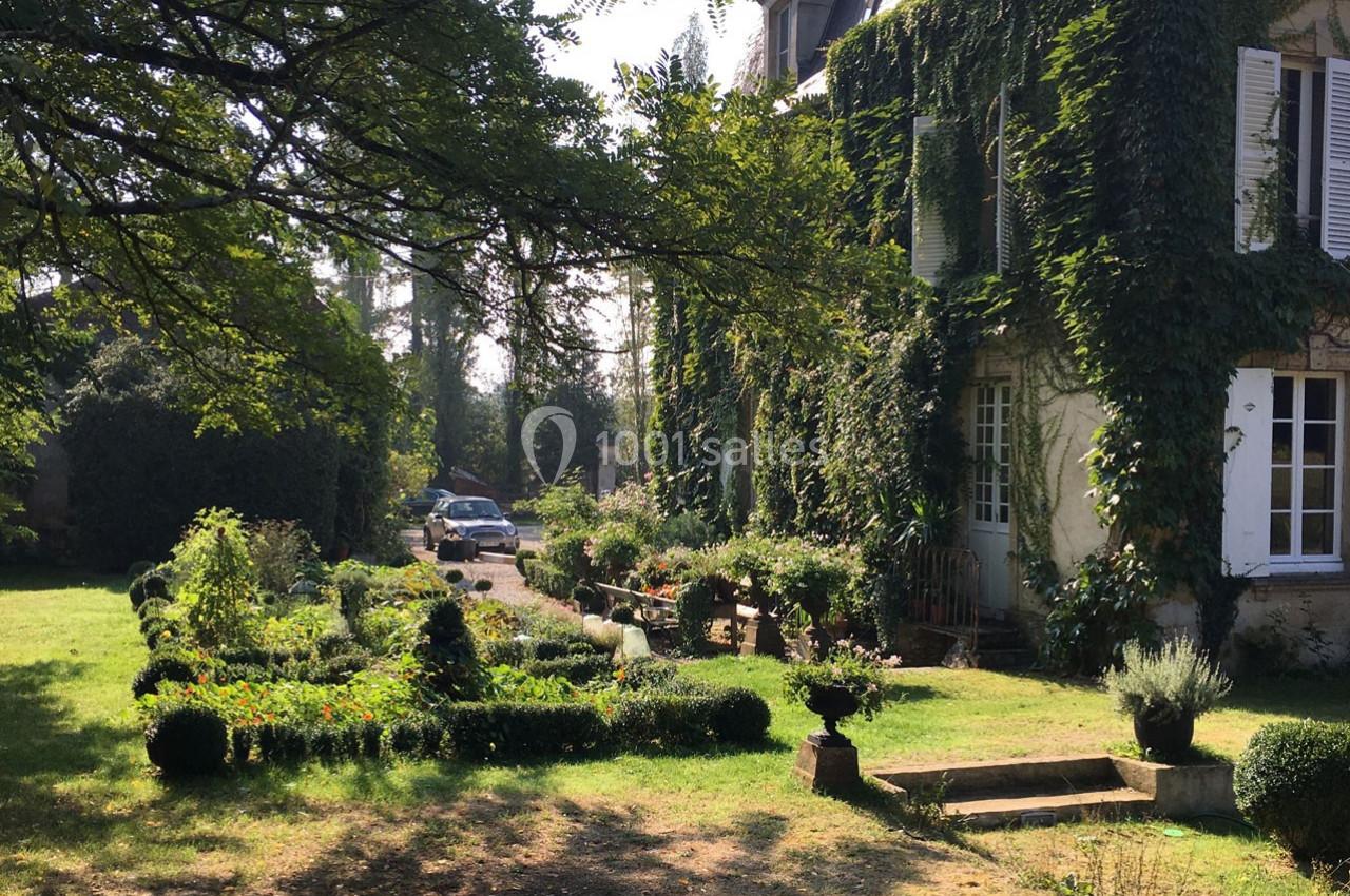 Maison ancienne couverte de lierre, entourée d'un jardin fleuri et arboré, avec une voiture garée à l'arrière-plan.