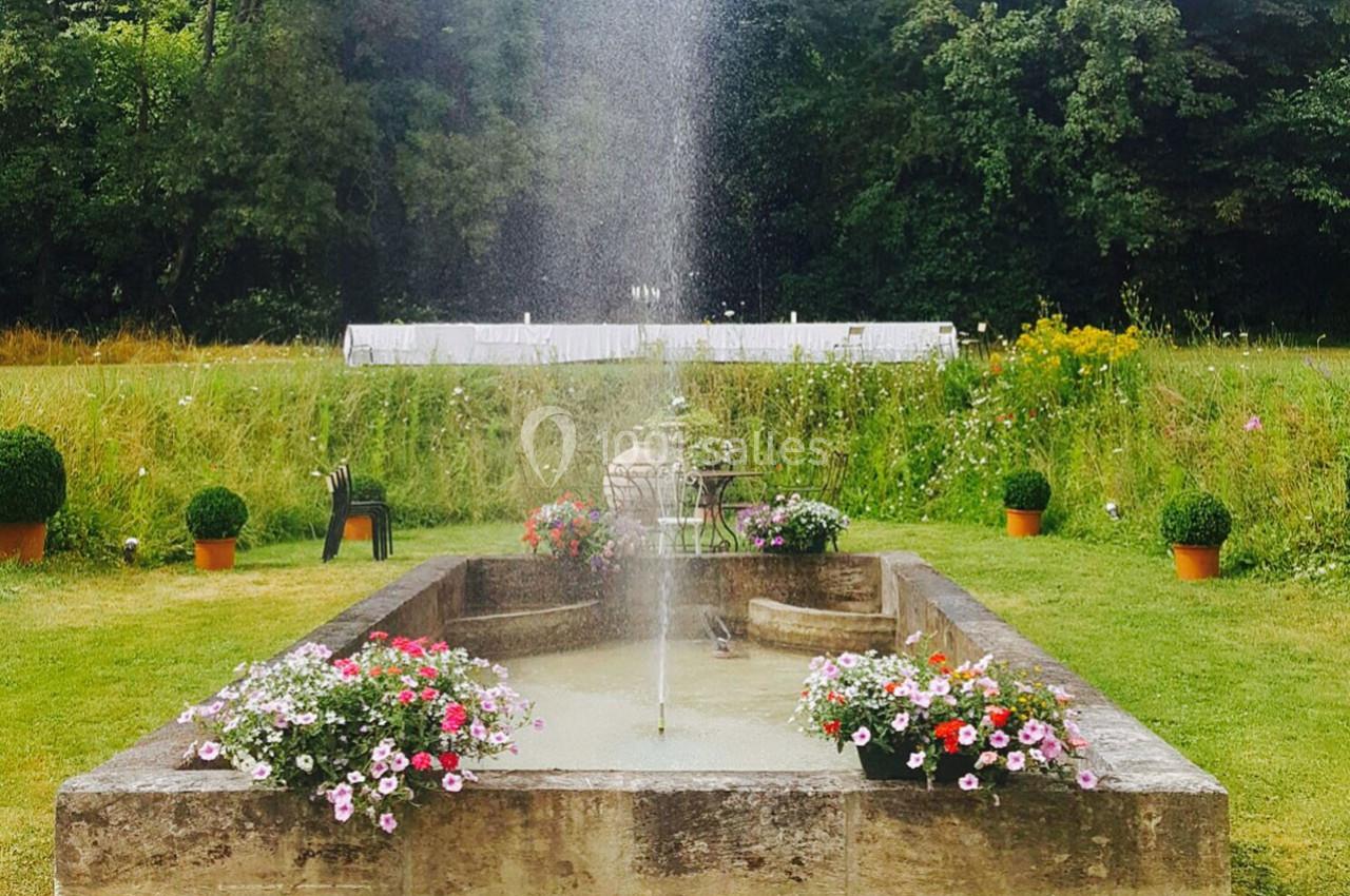 Fontaine centrale entourée de fleurs colorées dans un jardin verdoyant avec des arbres en arrière-plan.