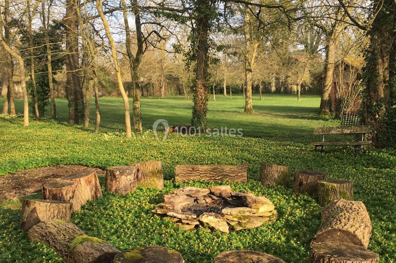 Rondins de bois disposés en cercle autour d'un foyer en pierre, dans une clairière verdoyante entourée d'arbres.