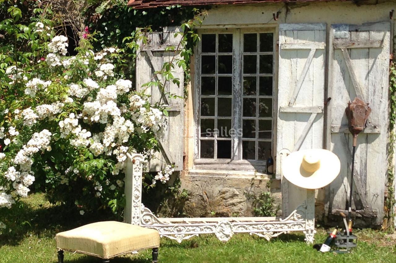 Petite maison en pierre avec volets en bois, entourée de fleurs blanches, un banc et un chapeau posés devant.
