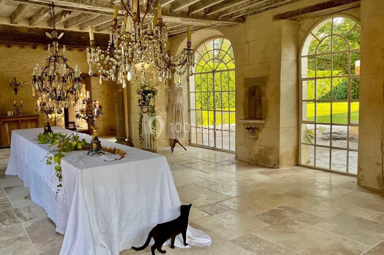 Salle lumineuse avec grandes fenêtres, table couverte d'une nappe blanche, lustres suspendus et un chat noir au sol.