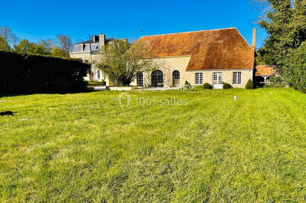 Grande maison en pierre avec toit en tuiles rouges, entourée d'un vaste jardin verdoyant sous un ciel bleu.