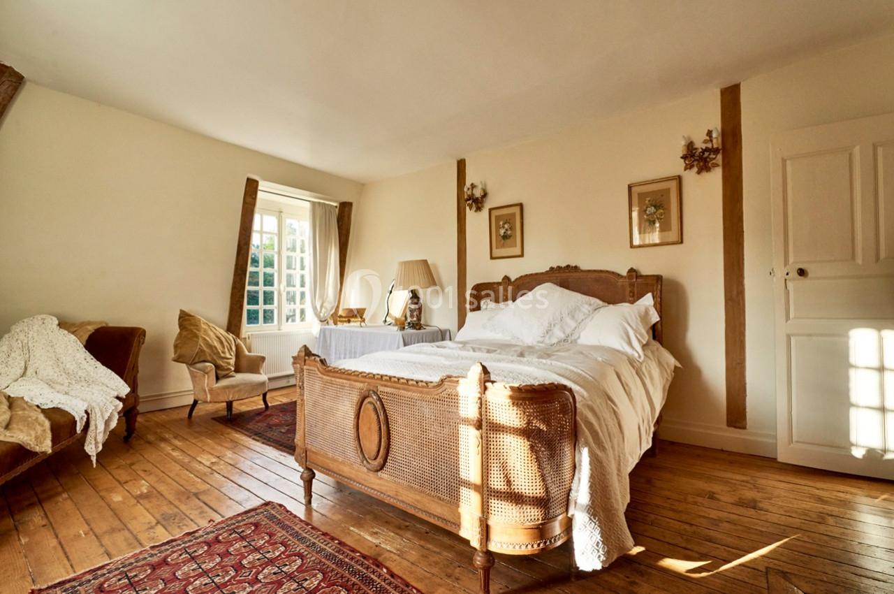 Chambre lumineuse avec lit ancien en bois, parquet, fauteuils, tapis et fenêtre donnant sur l'extérieur.