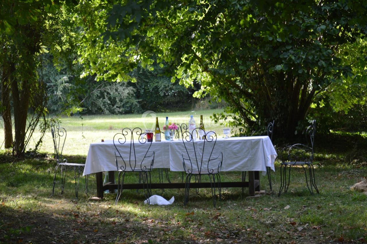 Table de jardin avec nappe blanche, chaises en fer forgé, bouteilles et verres, située sous des arbres dans un espace…