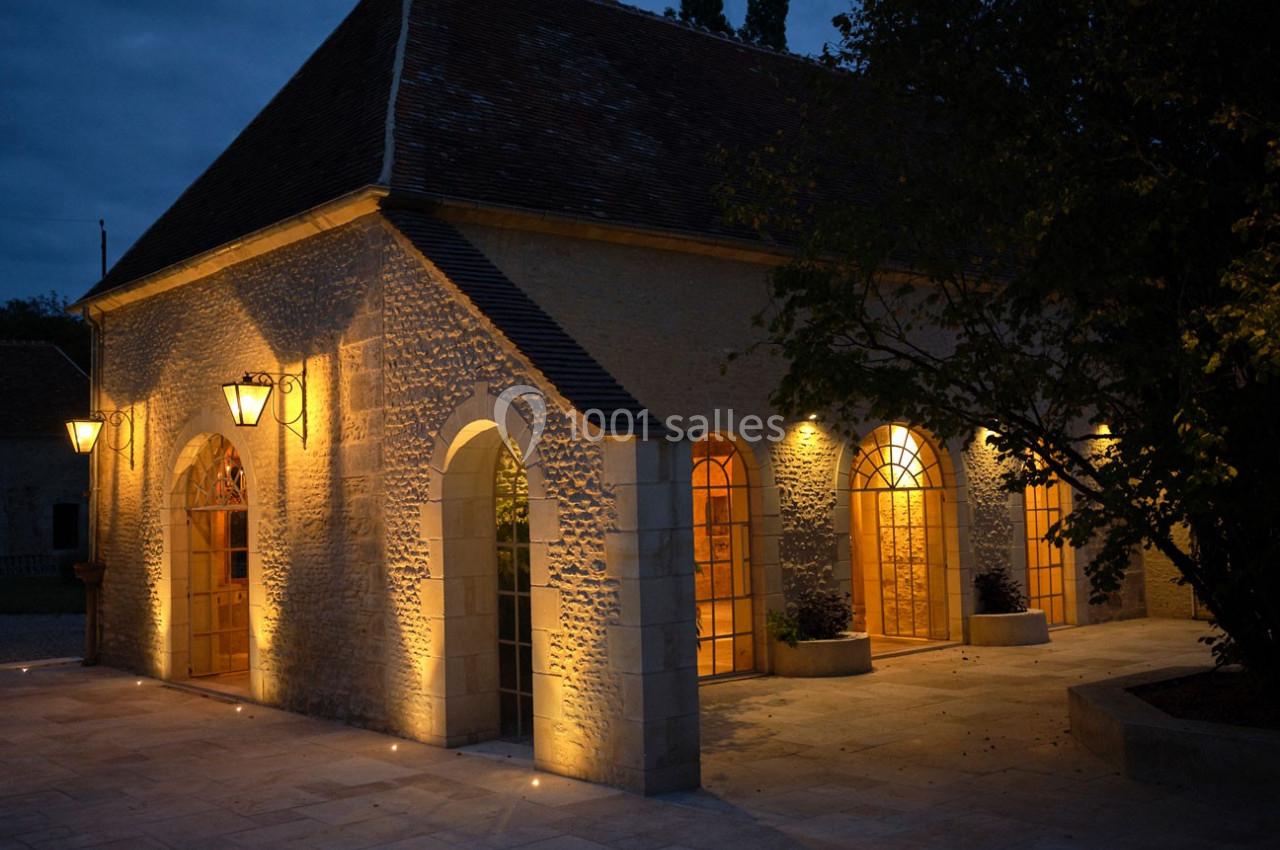 Bâtiment en pierre éclairé par des lampes extérieures, avec des arches vitrées, dans un cadre nocturne.
