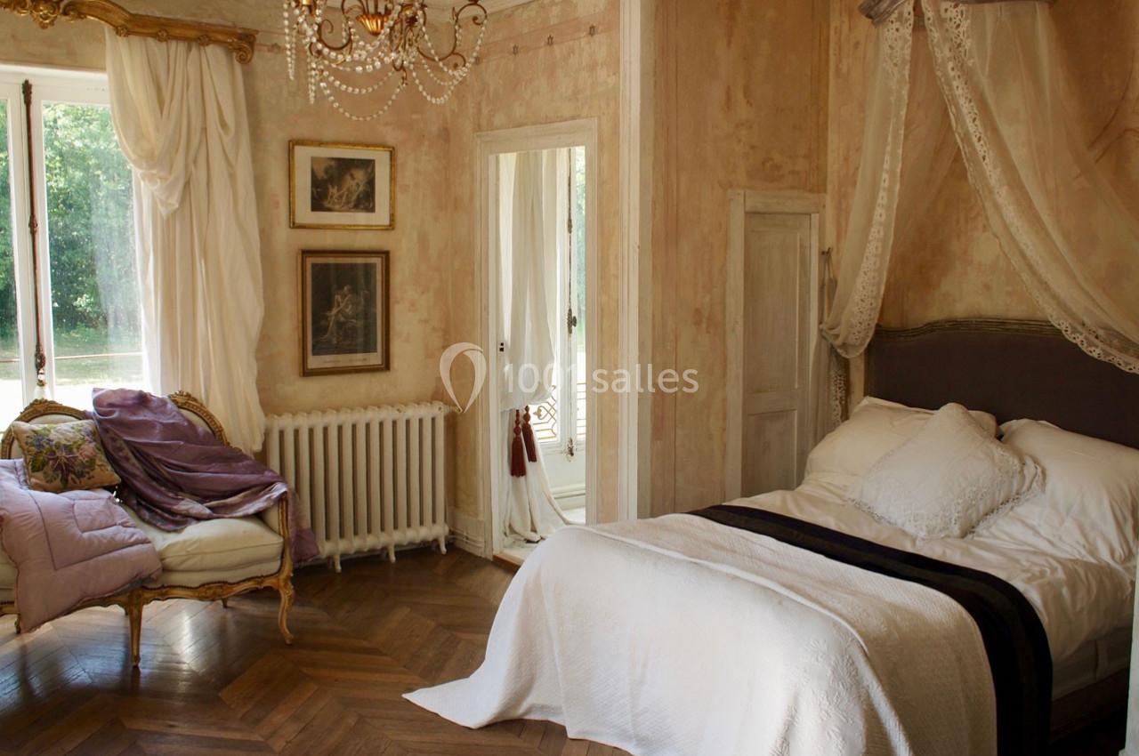 Chambre élégante avec lit à baldaquin, parquet en bois, fauteuil ancien et lumière naturelle entrant par une grande fenêtre.