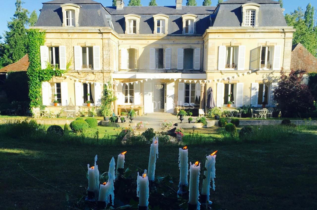 Façade d'un manoir en pierre entouré de verdure, avec des bougies allumées au premier plan.