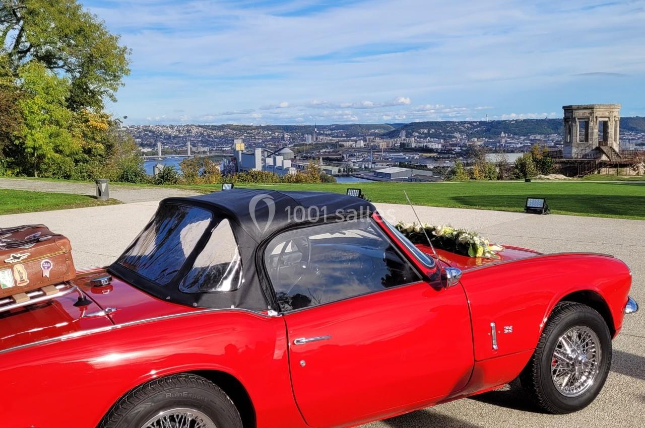 Voiture décapotable rouge avec valise ancienne sur le coffre, stationnée devant un panorama urbain et un monument.