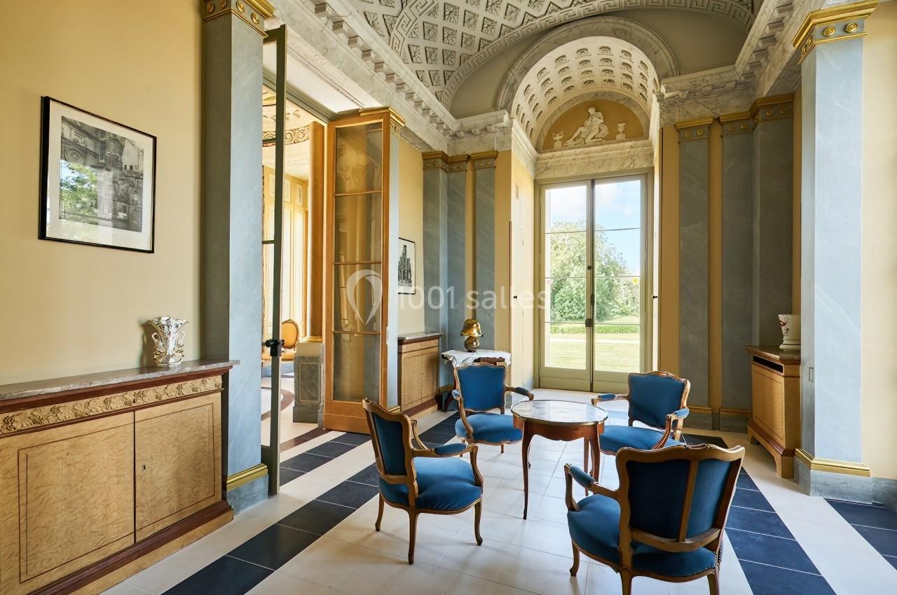 Salle élégante avec mobilier ancien, chaises bleues, table centrale, grandes fenêtres et décorations classiques.