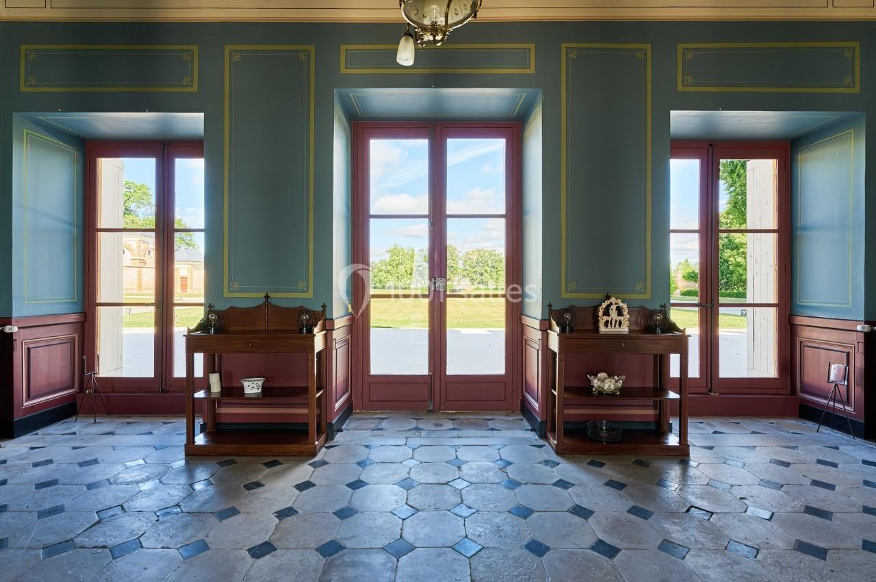 Salle lumineuse avec carrelage ancien, murs bleus ornés de moulures, et grandes portes vitrées donnant sur un jardin.