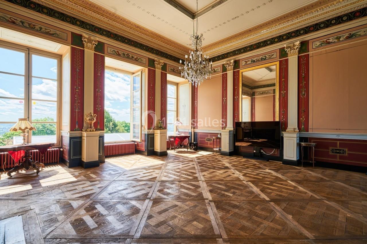 Salle élégante avec parquet en bois, murs ornés de moulures rouges et dorées, lustre central et grandes fenêtres lumineuses.