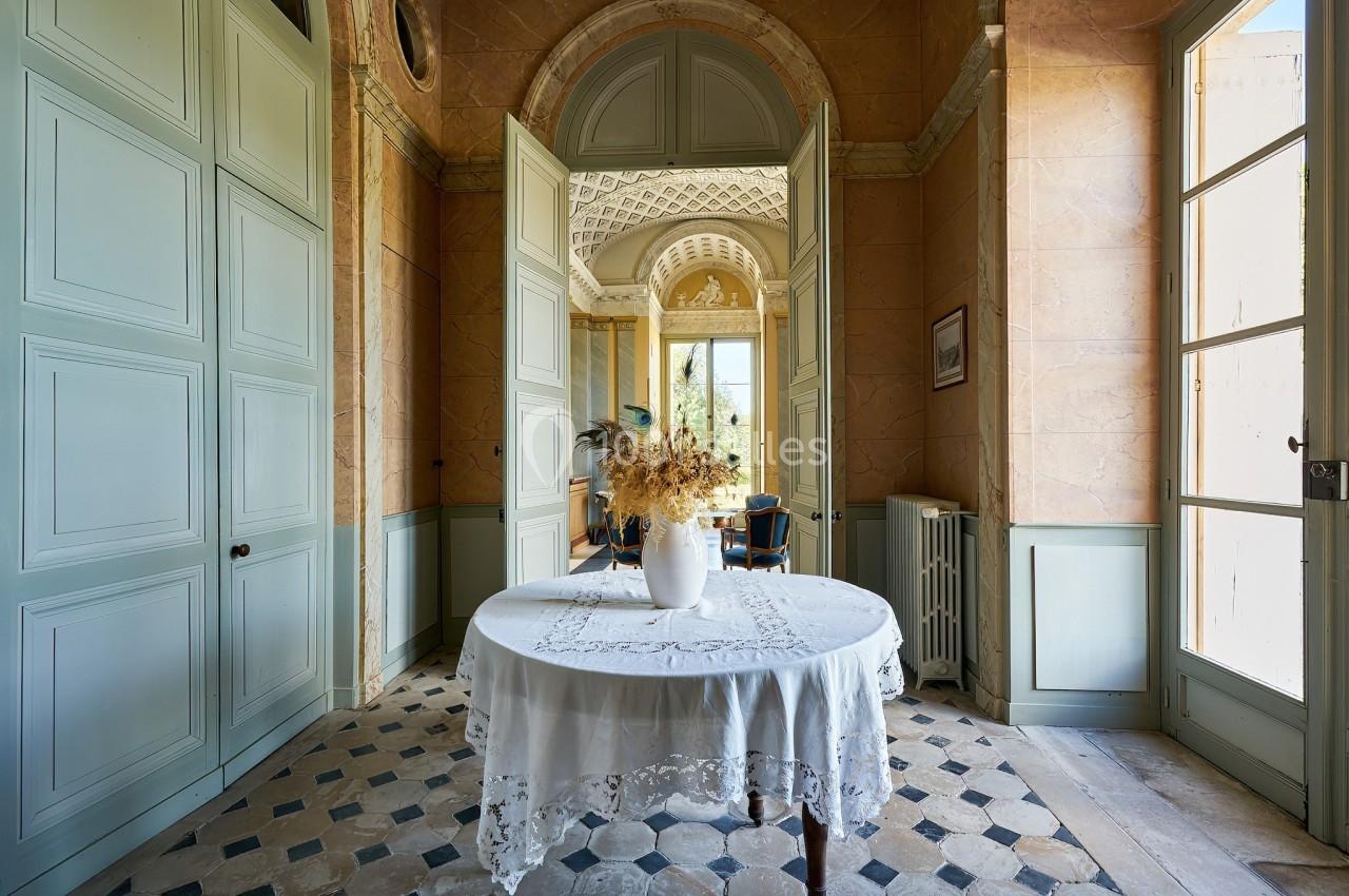 Intérieur lumineux avec table ronde ornée d'une nappe blanche et vase de fleurs séchées, portes ouvertes sur un salon.