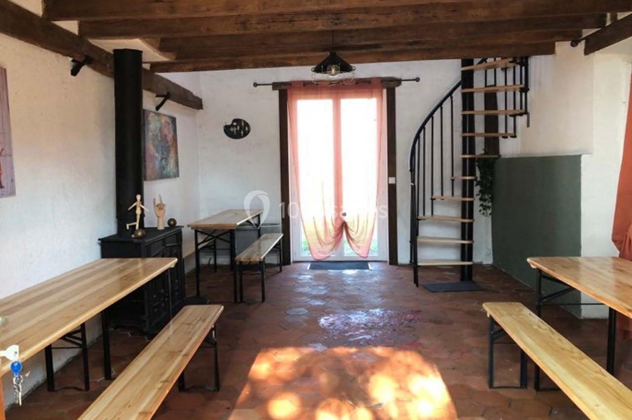 Salle lumineuse avec tables en bois, bancs, escalier en colimaçon, poutres apparentes et porte vitrée donnant sur l…